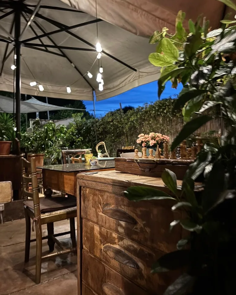 Il giardino all'imbrunire: ombrellone aperto, lampadine accese e un mobile di legno fra le piante.